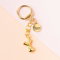 The-darling-effect's Y Bubble Letter Keychain Bag Charm features a shiny, abstract gold pendant and engraved round charm, making it a stylish personalized accessory—shown on a light peach and white background.