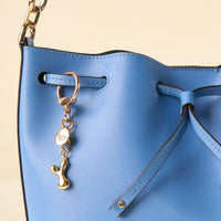 A close-up of a light blue leather handbag with a the-darling-effect Y Bubble Letter Keychain Bag Charm attached—a personalized round tag and small dog-shaped charm—plus gold hardware and a drawstring closure.