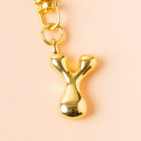 The Y Bubble Letter Keychain Bag Charm by the-darling-effect features a shiny gold, rounded "Y" with a gold clasp, shown on a light peach background—ideal for adding a personalized touch to any bag.