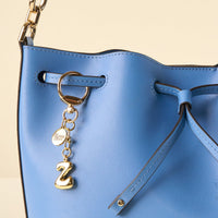 Close-up of a light blue leather handbag with a gold Z Bubble Letter Keychain Bag Charm by the-darling-effect, featuring a round charm and gold hardware. The bag has a drawstring closure and elegant gold accents.