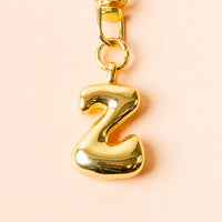 The-darling-effect Z Bubble Letter Keychain Bag Charm features a shiny gold bubble letter Z on a clasp, shown against a pale peach background—ideal for personalizing your bag.