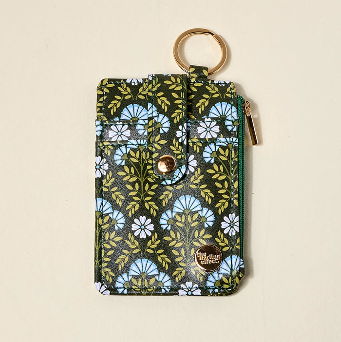 The Darling Effect’s Everbloom Forest Green Keychain Wallet features a floral pattern in blue, white, and green, gold snap button, green zipper, gold key ring, and round “Mighty Wonder” logo—perfect for stylish organization.