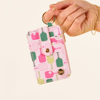 A manicured hand holds the Pickleball Party Keychain Wallet by the-darling-effect, featuring a pink, green, and yellow pickleball paddle and ball design, with a zipper, gold snap button, and keyring.