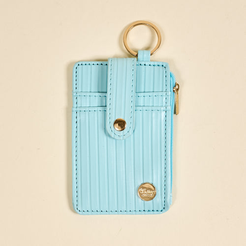 The Darling Effect Blue Ripple Ridge Keychain Wallet is a slim, light blue card holder with vertical stripes, a snap closure, gold keyring on top, right-side zipper, and a round gold logo at the bottom right.