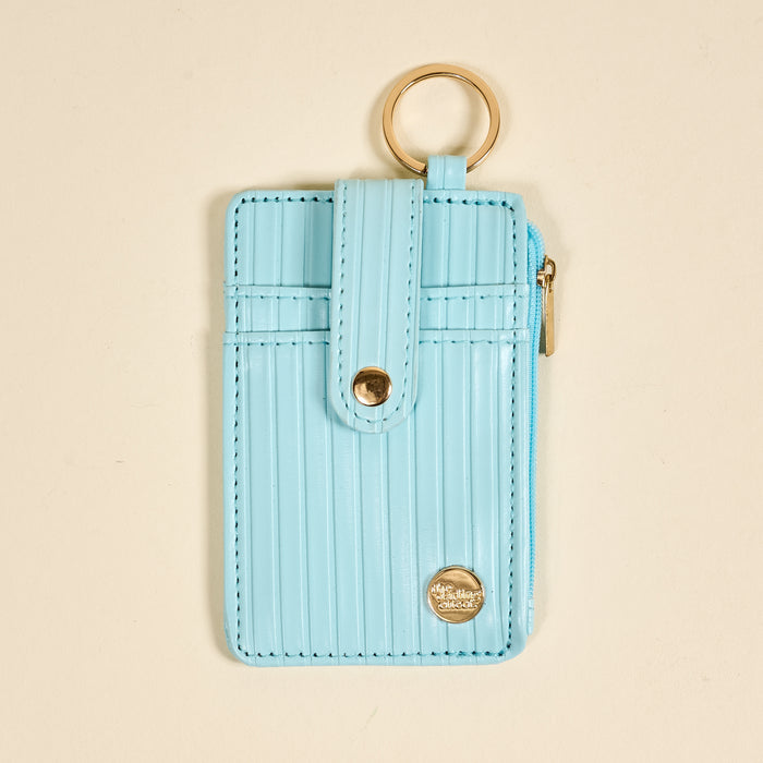 The Darling Effect Blue Ripple Ridge Keychain Wallet is a slim, light blue card holder with vertical stripes, a snap closure, gold keyring on top, right-side zipper, and a round gold logo at the bottom right.