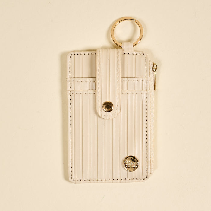 The Darling Effect's Oat Ripple Ridge Keychain Wallet, featuring beige vertical stripes, gold keyring, zippered coin pocket, and a snap-button flap closure, is displayed on a light neutral background.