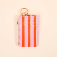 The Darling Effect’s Sicily Stripes Pink Keychain Wallet, with vertical pink and red stripes, features a zippered coin holder, card slots, and a gold keyring—shown on a light beige background.