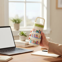 A hand holds The Darling Effect's Book Shelfie 6 oz Lil' Bitty Bottle, a petite white and cream vessel with a colorful bookshelf pattern. Laptop, notebooks, and plants rest on a wooden desk behind—ideal for travel.