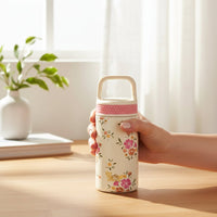 A hand holds The Darling Effect's Endless Daydream Cream 6 oz Lil' Bitty Bottle—a cream-colored flask with floral pattern, pink grip, and light beige handle—perfect for hydration on the go.