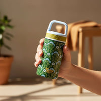 A hand holds the Everbloom Forest Green 6 oz Lil' Bitty Bottle by The Darling Effect, featuring a green pattern and blue-white lid. In the softly lit background, a wooden chair with brown cloth and a potted plant set the scene for portable hydration.