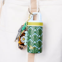 The Darling Effect’s Everbloom Forest Green 6 oz Lil' Bitty Bottle, with a yellow cap and green floral pattern, hangs from a beige bag by carabiner next to a beaded keychain—a stylish hand sanitizer ideal for travel.