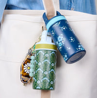 Two petite Lil' Bitty Bottles from The Darling Effect—one in Everbloom Forest Green—hang from a beige tote. A daisy beaded keychain with a tortoiseshell accent adds charm, with a denim shirt in the background. Perfect travel companions!.