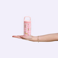 A hand with a bracelet holds the Oh So Fancy Pink 6 oz Lil' Bitty Bottle by The Darling Effect, a petite water bottle with floral and bow designs—perfect for on-the-go hydration—set against a plain light background.