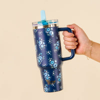 A hand holds the-darling-effect Endless Daydream Navy 40 oz Lifestyle Flip Straw Tumbler, featuring a blue floral pattern, clear lid, blue straw, and handle. Manicured nails and a gold bracelet add style against a beige background.