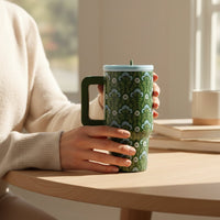 Wearing a cream sweater, a person sits at a wooden table holding The Darling Effect’s Everbloom Forest Green 20 oz Lifestyle Flip Straw Tumbler. Sunlight pours in, illuminating books and an insulated stainless steel cup on the table.