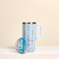 The Oh So Fancy Blue 20 oz Lifestyle Flip Straw Tumbler by the-darling-effect, featuring a tall, floral-patterned design and large handle, stands on a white surface with its clear blue flip straw lid in front. The background is beige.