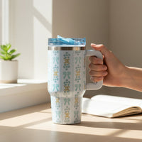 A hand holds the-darling-effect's Oh So Fancy Blue 20 oz Lifestyle Flip Straw Tumbler, featuring a floral pattern and blue lid, on a sunlit desk beside an open notebook and a small potted plant.
