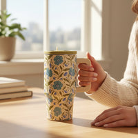 A person in a cream sweater holds The Darling Effect Poppin' Petals Aqua 20 oz Lifestyle Flip Straw Tumbler at a sunlit wooden table, with books and a potted plant by the window in the background.