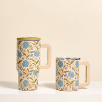 Two cream-colored mugs with blue floral designs and green leaves from The Darling Effect. One is the Poppin' Petals Aqua 20 oz Lifestyle Flip Straw Tumbler with lid; the other is shorter without a lid—both set on a white surface against a beige background.