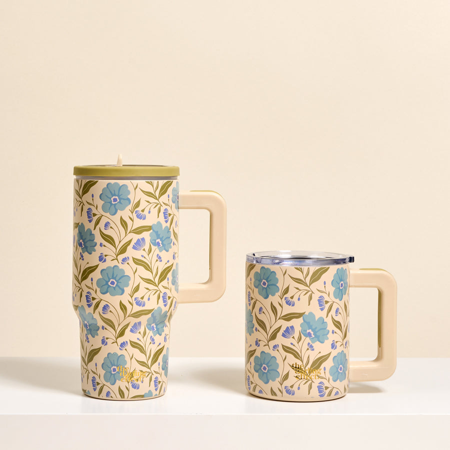 Two cream-colored mugs with blue floral designs and green leaves from The Darling Effect. One is the Poppin' Petals Aqua 20 oz Lifestyle Flip Straw Tumbler with lid; the other is shorter without a lid—both set on a white surface against a beige background.