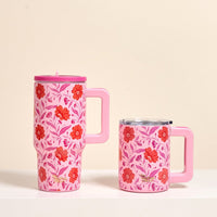 The Darling Effect's Poppin' Petals Hot Pink 20 oz Lifestyle Flip Straw Tumbler with a lid and floral pattern sits beside a shorter pink floral mug on a white surface against a beige background. Both mugs have handles and pink-red flower designs.