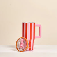 The Darling Effect Sicily Stripes Pink 20 oz Lifestyle Flip Straw Tumbler, featuring red and white stripes and a large pink handle, stands on a white surface with its clear lid beside it. The background is light beige.