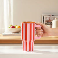 In a bright kitchen, a hand holds The Darling Effect Sicily Stripes Pink 20 oz Lifestyle Flip Straw Tumbler with a wooden lid. A bowl of fruit and kitchen items are softly lit in the background.