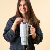 A smiling woman in a denim jacket holds the-darling-effect Oh So Fancy Blue 40 oz Lifestyle Flip Straw Tumbler with a floral pattern, set against a plain beige background.