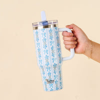 A hand holds the Oh So Fancy Blue 40 oz Lifestyle Flip Straw Tumbler by the-darling-effect, featuring a floral blue pattern, white handle, clear lid, leak-resistant flip straw, and logo against a plain beige background.