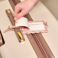 A hand attaches The Darling Effect Everbloom Dusty Rose Jetsetter Luggage Tag, featuring a floral pattern, privacy flap, and white insert for contact info, to a beige suitcase—an elegant travel accessory.