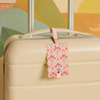 The Darling Effect’s Everbloom Dusty Rose Jetsetter Luggage Tag, featuring a scalloped edge, floral pattern, privacy flap, and pink strap, is attached to a beige suitcase for an elegant travel look.