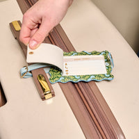A hand lifts the privacy flap of The Darling Effect Everbloom Forest Green Jetsetter Luggage Tag attached to a beige suitcase, revealing the contact info card—an elegant and functional travel accessory.