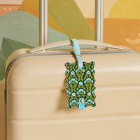 The Darling Effect’s Everbloom Forest Green Jetsetter Luggage Tag, featuring a privacy flap and light blue strap, attaches stylishly to a beige suitcase handle, set against a vibrant mural—perfect for chic travelers.