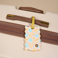 The Darling Effect’s Poppin' Petals Aqua Jetsetter Luggage Tag, with its floral design and privacy flap, hangs from a brown-handle cream suitcase—making it the perfect travel accessory.