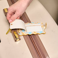 A hand lifts the privacy flap of The Darling Effect’s Poppin' Petals Aqua Jetsetter Luggage Tag—attached to a beige suitcase—revealing a card below for name, address, and phone number.