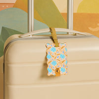 The Darling Effect's Poppin' Petals Aqua Jetsetter Luggage Tag—a stylish floral accessory with blue flowers and a wavy edge—adorns a cream hard-shell suitcase with a gold handle, set against a colorful abstract background.