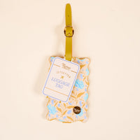 The Darling Effect’s Poppin' Petals Aqua Jetsetter Luggage Tag features a yellow strap, scalloped edges, a yellow and blue floral design, privacy flap, and “JETSETTER LUGGAGE TAG” label on a light beige background.