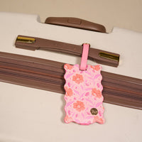 The Darling Effect’s Poppin' Petals Hot Pink Jetsetter Luggage Tag, featuring scalloped edges and a privacy flap, is attached to the handle of a white suitcase with brown accents for a charming touch.