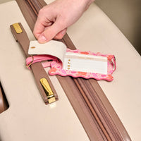 A hand lifts the privacy flap on The Darling Effect’s Poppin' Petals Hot Pink Jetsetter Luggage Tag, attached to a beige suitcase, revealing a name and address card inside this chic floral travel accessory.