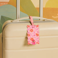 A cream-colored hard-shell suitcase with The Darling Effect’s Poppin’ Petals Hot Pink Jetsetter Luggage Tag on the handle sits in front of a vibrant abstract mural.