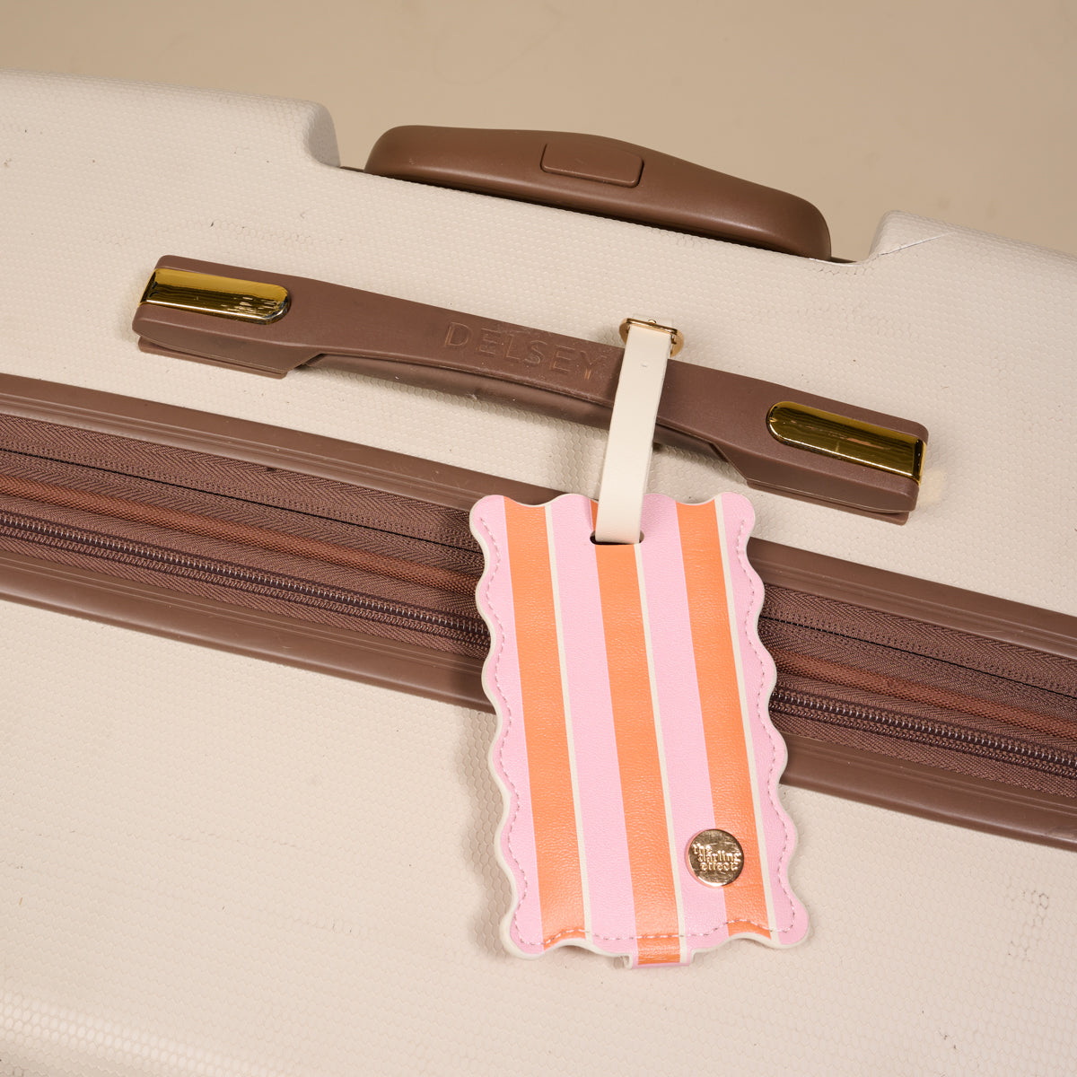 The Sicily Stripes Pink Jetsetter Luggage Tag by The Darling Effect, featuring pink and orange stripes with a scalloped edge and privacy flap, hangs from a tan Delsey suitcase as a stylish travel accessory.