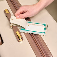 A hand lifts the privacy flap of The Darling Effect’s Sicily Stripes Teal Jetsetter Luggage Tag on a beige suitcase, revealing icons for name, address, and contact info—an essential travel accessory for safeguarding personal details.