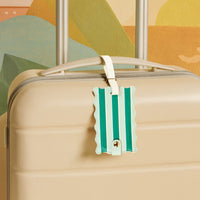 The Darling Effect’s Sicily Stripes Teal Jetsetter Luggage Tag, with teal and light teal stripes plus a privacy flap, adds style to a beige suitcase handle against a colorful abstract background—a chic travel accessory.