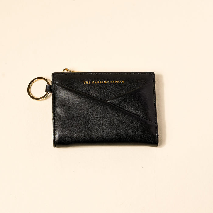 The Darling Effect’s Black Luxe Pocket Wallet, made from black vegan leather with a gold zipper and key ring, features the brand name embossed in gold on the front and is shown on a light beige background.