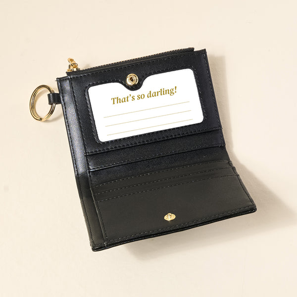 The Darling Effect Black Luxe Pocket Wallet, made of vegan leather with a gold keyring, is open to show card slots and a “That’s so darling!” card in gold. This compact wallet is photographed against a beige background.
