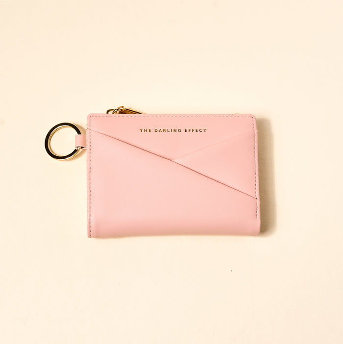 The Blush Luxe Pocket Wallet by The Darling Effect is a light pink vegan leather compact wallet with a gold keyring and zipper, featuring “THE DARLING EFFECT” embossed in gold on the front, shown against a beige background.