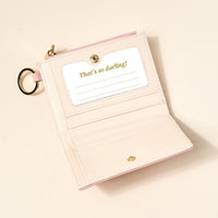 The Darling Effect’s Blush Luxe Pocket Wallet, a pale pink vegan leather accessory, features card slots, a clear ID window with a “That’s so darling!” insert, and a gold keyring, shown open on a light surface.