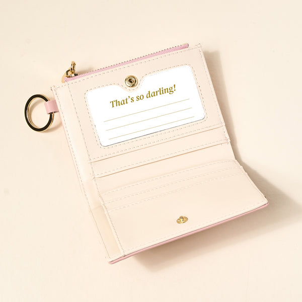 The Darling Effect’s Blush Luxe Pocket Wallet, a pale pink vegan leather accessory, features card slots, a clear ID window with a “That’s so darling!” insert, and a gold keyring, shown open on a light surface.