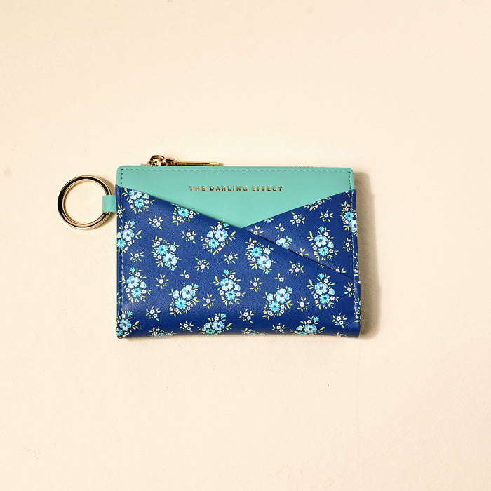 The Darling Effect Endless Daydream Navy Luxe Pocket Wallet features a blue floral pattern, light blue flap with gold "THE DARLING EFFECT" text, gold zipper, key ring, and vegan leather. Shown on a cream background.