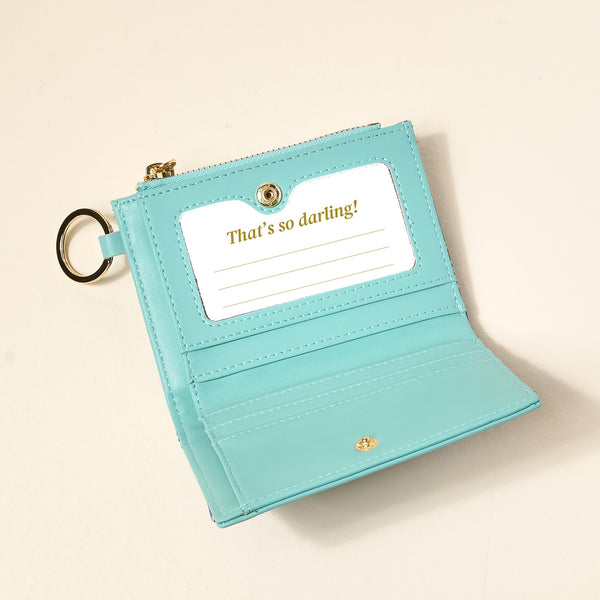 The Darling Effect's Endless Daydream Navy Luxe Pocket Wallet, made of vegan leather, is open to show a card slot with a card that says "Thats so darling!" in gold and three blank lines, placed on a light surface.